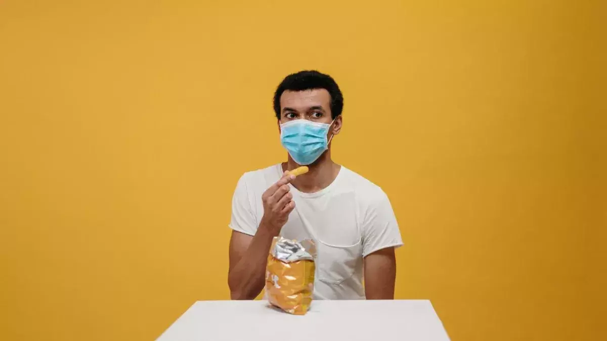 Ein Mann mit einer Tüte Chips und Maske vor gelbem Hintergrund