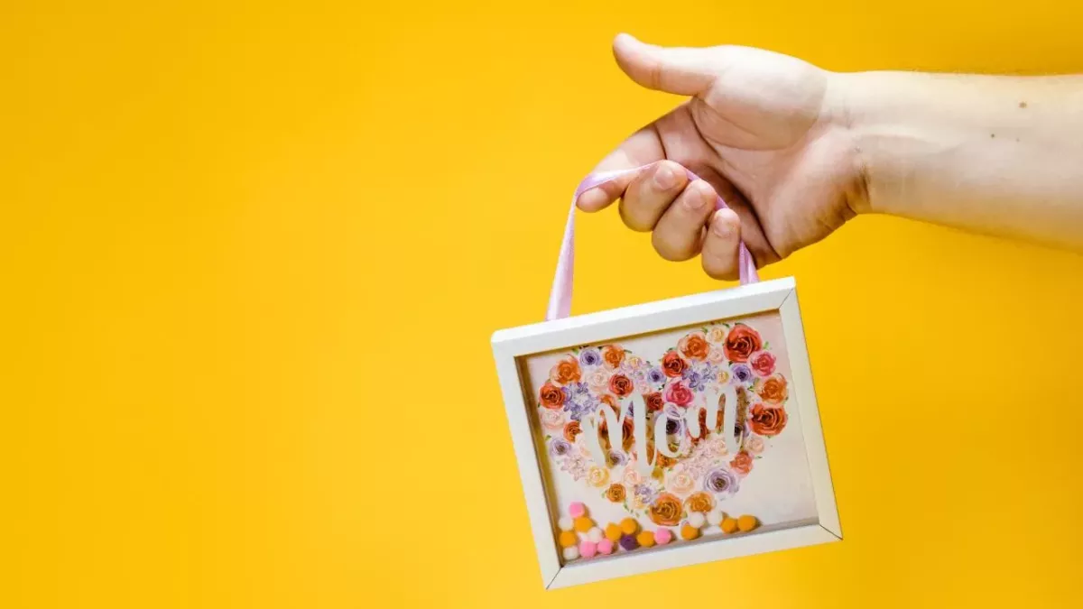 Eine Hand mit kleinem Bilderrahmen, der den Schriftzug "Mom" trägt, vor gelbem Hintergrund