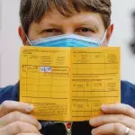 Ein Mann mit Mund-Nasen-Schutz hält einen gelben Impfpass in den Händen