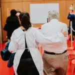 Frau führt älteren Mann zum Platz, an dem er seine Spritze bekommt