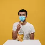 Ein Mann mit einer Tüte Chips und Maske vor gelbem Hintergrund