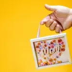 Eine Hand mit kleinem Bilderrahmen, der den Schriftzug "Mom" trägt, vor gelbem Hintergrund