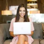 Eine lächelnde Frau sitzt mit einem Laptop auf dem Schoß auf einer Couch