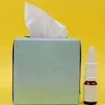 Eine Packung Taschentücher und ein Nasenspray vor gelbem Hintergrund