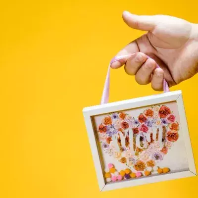 Eine Hand mit kleinem Bilderrahmen, der den Schriftzug "Mom" trägt, vor gelbem Hintergrund